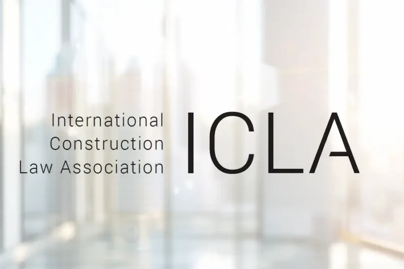 Logo of the International Construction Law Association (ICLA) displayed in front of a bright office window with soft sunlight and a blurred urban skyline in the background.