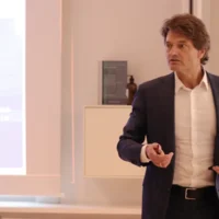 A professionally dressed man, Dr. Wolfgang Breyer speaks in front of a projected slide during an ICLA event, gesturing while holding a presentation remote. A blurred screen and audience members are visible in the background.