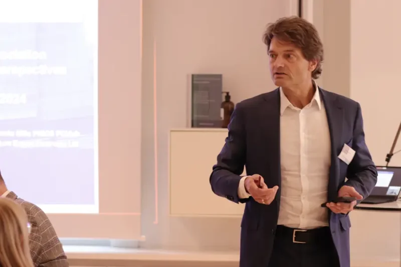 A professionally dressed man, Dr. Wolfgang Breyer speaks in front of a projected slide during an ICLA event, gesturing while holding a presentation remote. A blurred screen and audience members are visible in the background.