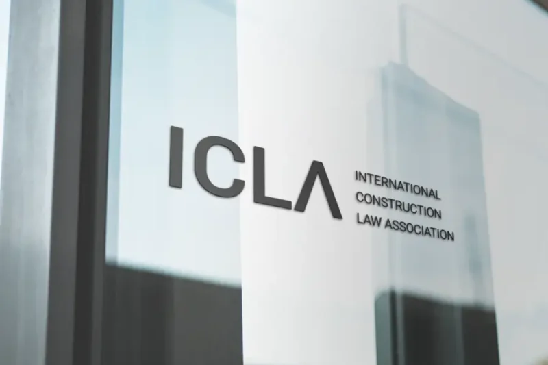 Logo of the International Construction Law Association (ICLA) applied to a glass office door or window, with urban reflections faintly visible. The black letters contrast with the bright, modern surroundings, conveying a professional and international atmosphere.